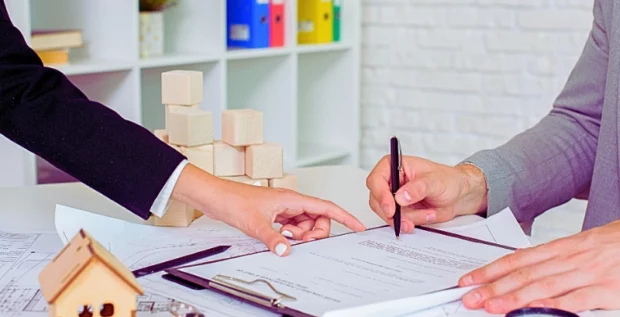 Woman pointing where the other woman to sign the document with wooden house, calculator. keys and magnifying glass on the desk. Quitclaim Deeds