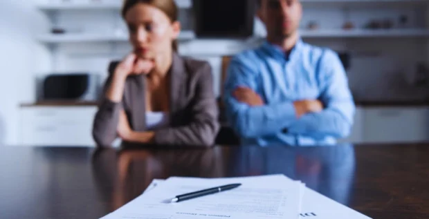 A man and a woman sitting at a table with a stack of documents in front of them. On top of the papers, there are two gold wedding rings and a pen. Divide a business in an Illinois divorce